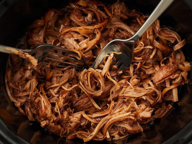 Slow cooker pulled pork being shredded with two forks, showing tender, juicy meat